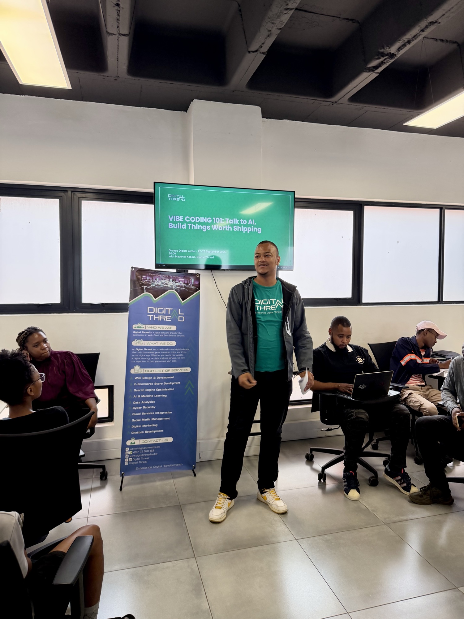 Maverick Kabelo presenting in front of the Vibe Coding 101 title slide, beside a Digital Thread banner, with participants on laptops